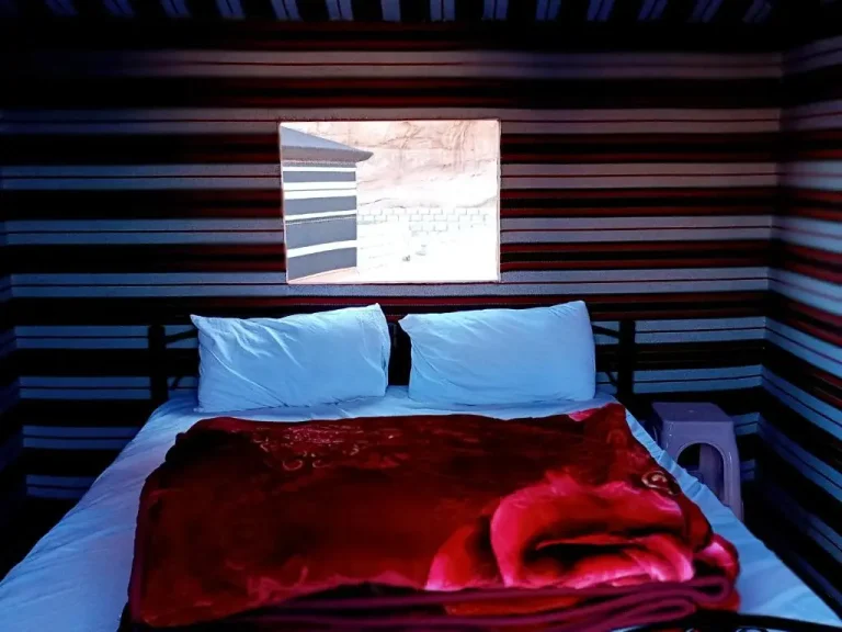 A cozy double bed inside a traditional Bedouin tent at Salem Camp in Wadi Rum, with a colorful red blanket and striped walls.
